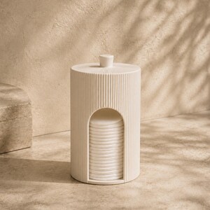 Può includere: Un dispenser cilindrico per dischetti di cotone color bianco sporco con una texture a coste verticali e un piccolo pomello sul coperchio. Il dispenser presenta un'apertura ad arco alla base, che rivela una pila di dischetti bianchi.