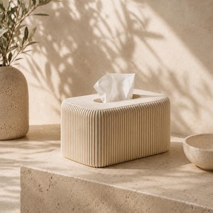Puede incluir: Una caja para pañuelos rectangular con una textura acanalada vertical en color crema sólido. La caja presenta una superficie superior lisa y redondeada con una abertura ovalada central de la que sobresale un pañuelo blanco.