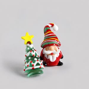 Può includere: Un set di statuine in vetro composto da un albero di Natale verde con ghirlanda bianca a zigzag, puntini rossi e una stella gialla in cima, insieme a uno gnomo in vetro con cappello a righe rosse, verdi e bianche, barba bianca e cappotto rosso.
