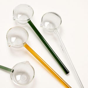 May include: Four glass ladles with clear, round bowls and small pouring spouts. Each ladle features a long, slender handle in a different color: one dark green, one amber, one sage green, and one clear glass, all arranged on a plain white surface.