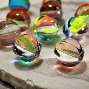 May include: A collection of spherical glass beads featuring swirling patterns of vibrant colours including lime green, orange, blue, and translucent pink. Each bead has a smooth, polished surface that reflects light, creating a glossy appearance.