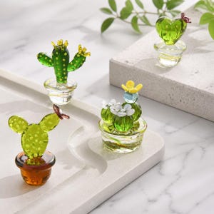 May include: Four glass cactus figurines in small pots. One is a tall green cactus with yellow accents, one is a heart-shaped green cactus, one is a round green cactus with white and yellow flowers, and one is a flat green cactus in a brown pot.