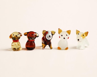 May include: A set of five miniature glass dog figurines arranged in a row. The collection includes a translucent amber dog with a red collar, a dark brown dog with a red collar, a brown and white spotted dog, a white dog with orange ears, and a white dog.