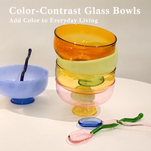 Könnte beinhalten: Ein Stapel durchscheinender Glasschalen in Bernstein, Gelb, Grün und Rosa neben einer hellblauen Schale mit einem dunkelblauen Wellenlöffel. Ein weiterer rosa-grüner Wellenlöffel liegt daneben. Text: Color-Contrast Glass Bowls Add Color to Everyday Living.