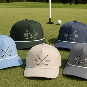 May include: Five structured baseball caps in light blue, dark green, navy blue, beige, and gray. Each cap features a white rope detail across the brim and a graphic print of two crossed golf clubs, a golf ball, and the text EST. 1996.