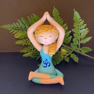 May include: A small figurine of a person in a blue yoga outfit with blonde hair sitting in a meditation pose with their hands raised above their head. The figurine has an Om symbol on the front of their outfit.