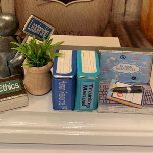 May include: A miniature figurine of a person holding a sign that says "Leadership". The figurine is standing next to a potted plant, a book titled "Ethics", two books titled "Human Resources 101" and "Training Manual", and a laptop with a screen that says "Human Resources".