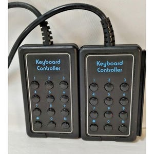 May include: Two black rectangular keyboard controller units, each featuring a numeric keypad with twelve circular buttons labeled 0 through 9, an asterisk, and a pound sign. The text Keyboard Controller is printed in blue above the buttons.