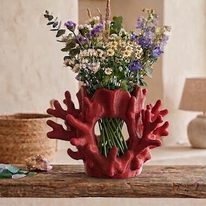 Può includere: Un vaso rosso intenso a forma di corallo con un'apertura circolare centrale, contenente un vivace bouquet di fiori selvatici viola, bianchi e verdi. Il vaso presenta complesse texture ramificate che imitano l'aspetto del corallo marino naturale.