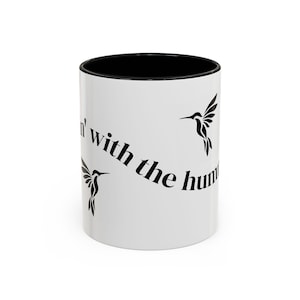 May include: White ceramic mug with a black interior and the phrase "Flyin' with the hum" in black script. The mug features three black hummingbird silhouettes, creating a decorative design.
