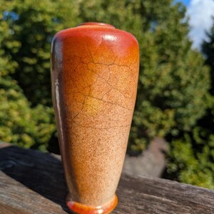 Puede incluir: Un jarrón de cerámica con una silueta cónica y acabado brillante. La superficie presenta un degradado de colores cálidos, pasando de un rojo anaranjado intenso en la parte superior a un tono beige moteado más claro en la base, con esmalte craquelado.