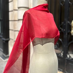 May include: A vibrant red scarf made of a lightweight, semi-sheer fabric draped over a beige mannequin. The scarf features a rectangular shape with finished edges, showcasing a smooth texture and a consistent, solid red color throughout the material.
