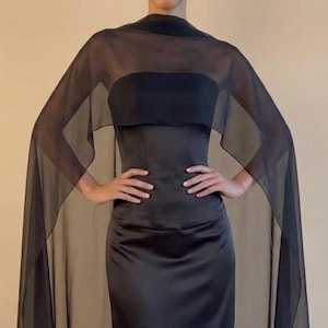 Peut inclure: Une robe longue en satin noir dotée d'un corsage sans bretelles et d'une cape transparente et légère en mousseline noire qui recouvre les épaules et descend sur les côtés, créant une silhouette fluide avec une finition lisse et unie.