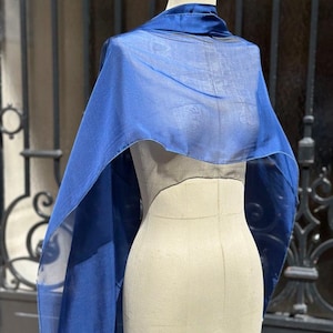 Peut inclure: Un châle bleu profond et transparent drapé sur un torse de mannequin beige. Le tissu est léger et translucide, présentant une texture lisse et une couleur bleue unie et constante qui tombe des épaules en longs panneaux fluides.