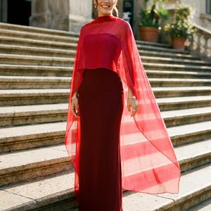 May include: A floor-length, dark red strapless gown featuring a fitted silhouette and a sheer, bright red cape overlay that drapes over the shoulders and extends down the back, creating a layered effect with contrasting shades of red fabric.