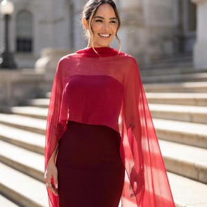 Op de afbeelding: Een diep bordeauxrode strapless jurk gecombineerd met een doorschijnende, felrode capelet die over de schouders valt. De jurk heeft een nauwsluitend silhouet en een rechte halslijn, terwijl de lichtgewicht capelet een vloeiende laag toevoegt.
