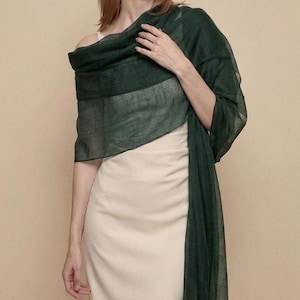 May include: A long, rectangular, dark forest green scarf made of a lightweight, semi-sheer fabric draped over a solid cream-colored sleeveless dress. The scarf features a fine, woven texture and hangs loosely over the shoulders and down the side.