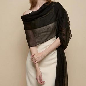 May include: A sheer, rectangular black shawl draped over the shoulders, featuring a lightweight, semi-transparent fabric with a fine weave. The shawl is worn over a solid, sleeveless cream-colored dress with a simple, straight neckline.