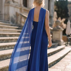 May include: A floor-length royal blue gown featuring a sleeveless bodice with a deep open back and long, flowing sheer chiffon cape panels attached at the shoulders that drape down the back to create a dramatic, elegant silhouette.