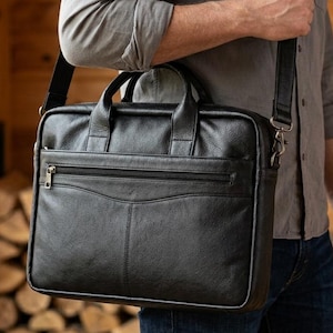 May include: A black leather briefcase featuring a rectangular shape, two top handles, and an adjustable shoulder strap. The front includes a horizontal zipped pocket with a metallic zip pull and a curved stitched seam detail across the centre.