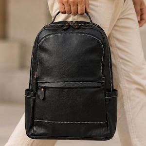 May include: A black leather backpack featuring a top handle, a main zippered compartment, and a smaller front zippered pocket. The bag includes side pockets and is accented with visible white stitching along the seams and dark metal hardware.