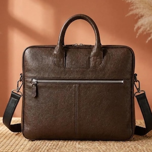 May include: A dark brown leather briefcase featuring two top handles, a horizontal zipped front pocket with a silver-toned metal zip, and a detachable black shoulder strap with metal hardware clips attached to the sides of the bag.