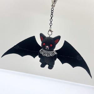 May include: A black bat keychain featuring a textured body, large red eyes, pointed ears with red inner details, and two white fangs. The bat wears a white lace collar and has wide, outstretched black wings, attached to a silver metal chain and clasp.