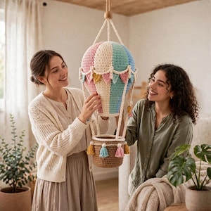 Può includere: Una decorazione a mongolfiera sospesa realizzata in filo all'uncinetto nei toni pastello del rosa, crema e blu. Il pallone presenta un bordo smerlato decorativo ed è sospeso sopra un cestino all'uncinetto marrone decorato con nappe colorate.
