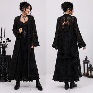 May include: A long, black, open-front crochet duster cardigan featuring wide sleeves, a scalloped hem, and a large circular floral motif on the back. The garment is worn over a black dress and paired with black ankle boots, creating a layered look.