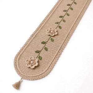 May include: A beige crochet table runner featuring a central vine pattern with green leaves and two dimensional flowers. Each flower is accented with a white pearl bead at the center. The runner has a rounded end finished with a small beige tassel.