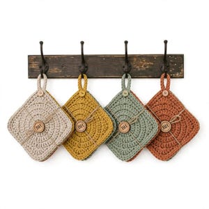 May include: Four square crochet potholders in beige, mustard yellow, sage green, and terracotta orange, each featuring a small wooden button and a circular tag with the letters BH, hanging from a dark brown distressed wooden rack with four hooks.