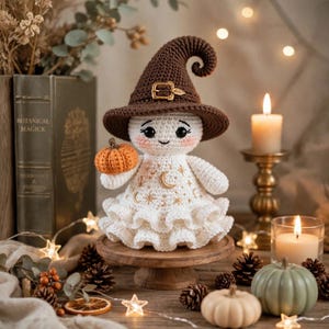 May include: A crocheted ghost figurine wearing a brown pointed hat with a gold buckle. The white ghost has embroidered gold moon and star patterns on its dress, a ruffled hem, and holds a small orange crocheted pumpkin in its right hand.