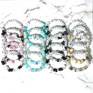 May include: A collection of sixteen circular beaded bracelets featuring white letter beads spelling out song titles. The bracelets are arranged in four columns, showcasing various color schemes including pink, black, teal, blue, and metallic gold tones.