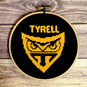 May include: A black embroidery hoop with a yellow embroidered design of a stylised owl with the word "TYRELL" above it.