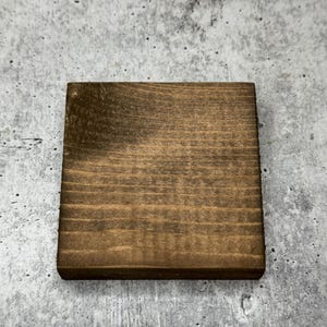 May include: A square wooden coaster with a dark brown stain. The coaster has a natural wood grain pattern.