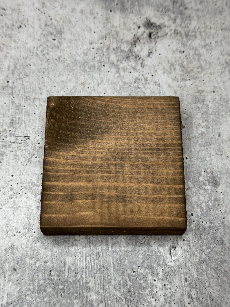 May include: A square wooden coaster with a dark brown stain. The coaster has a natural wood grain pattern.