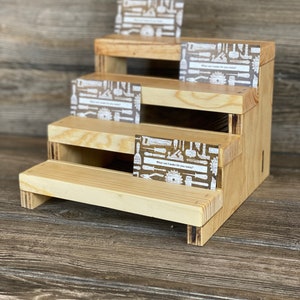 May include: A wooden tiered display stand with four shelves. Each shelf has a small card with a white background and a pattern of tools in black. The text on the card reads "What can I make for you today?"
