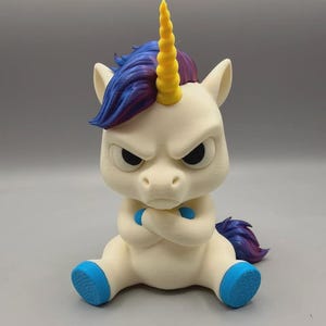 May include: A cream-colored unicorn figurine with a grumpy facial expression and crossed arms. It features a bright yellow spiral horn, a blue and purple mane, a matching blue and purple tail, and bright blue hooves on its seated legs.