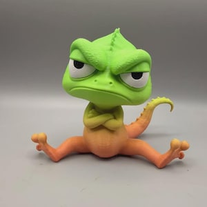 May include: A 3D printed chameleon figurine with a large head, furrowed brow, and crossed arms. The creature features a bright green upper body and head, transitioning into an orange lower body and limbs, with a curved tail extending behind.
