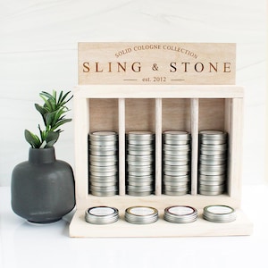 May include: A wooden display stand with the text "SOLID COLOGNE COLLECTION SLING & STONE est. 2012" on the top. The stand has four compartments filled with stacks of small, round, silver tins. There are four smaller tins on the bottom shelf, each with a different colored label.