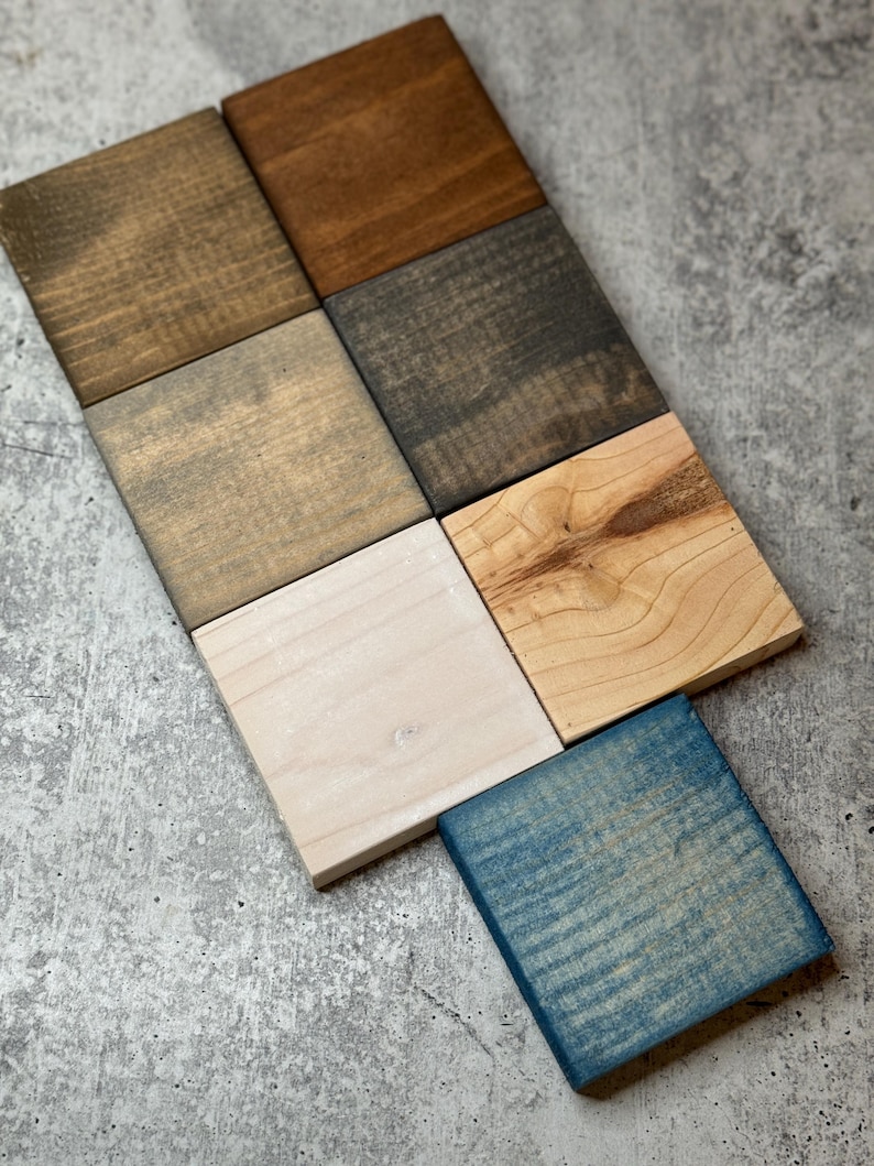 May include: Eight wood samples stained in different colors, including natural wood, brown, gray, white, and blue. The samples are arranged in a staggered pattern on a gray concrete surface.