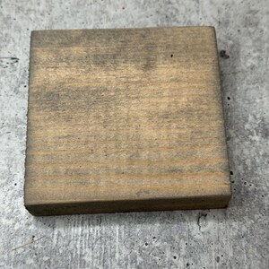 May include: A square piece of wood with a natural wood grain and a light brown colour. The wood has a distressed look with some darker areas.