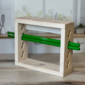 May include: Wooden drumstick holder with a natural finish. The holder has a rectangular shape with rounded edges and features a series of holes on the sides. Two green drumsticks are resting inside the holder.