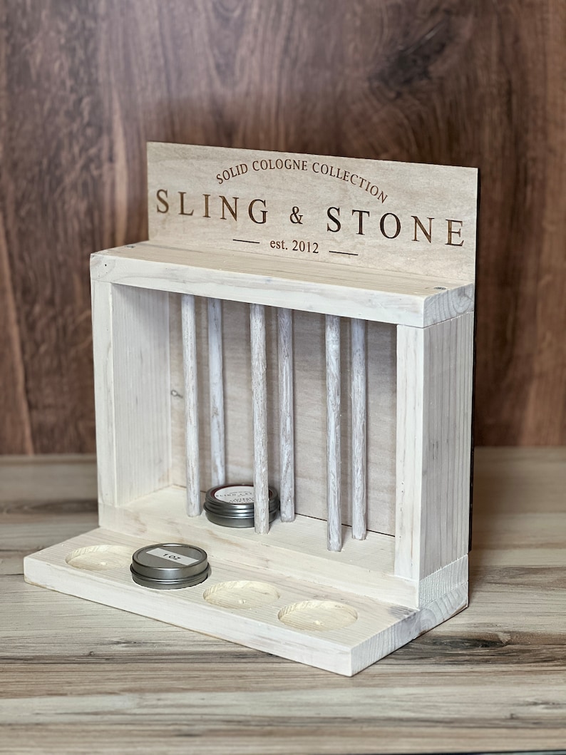 May include: White wooden display stand with a sign that reads "Solid Cologne Collection Sling & Stone est. 2012". The stand has a shelf with four round holes and a vertical section with slots for holding small round containers.