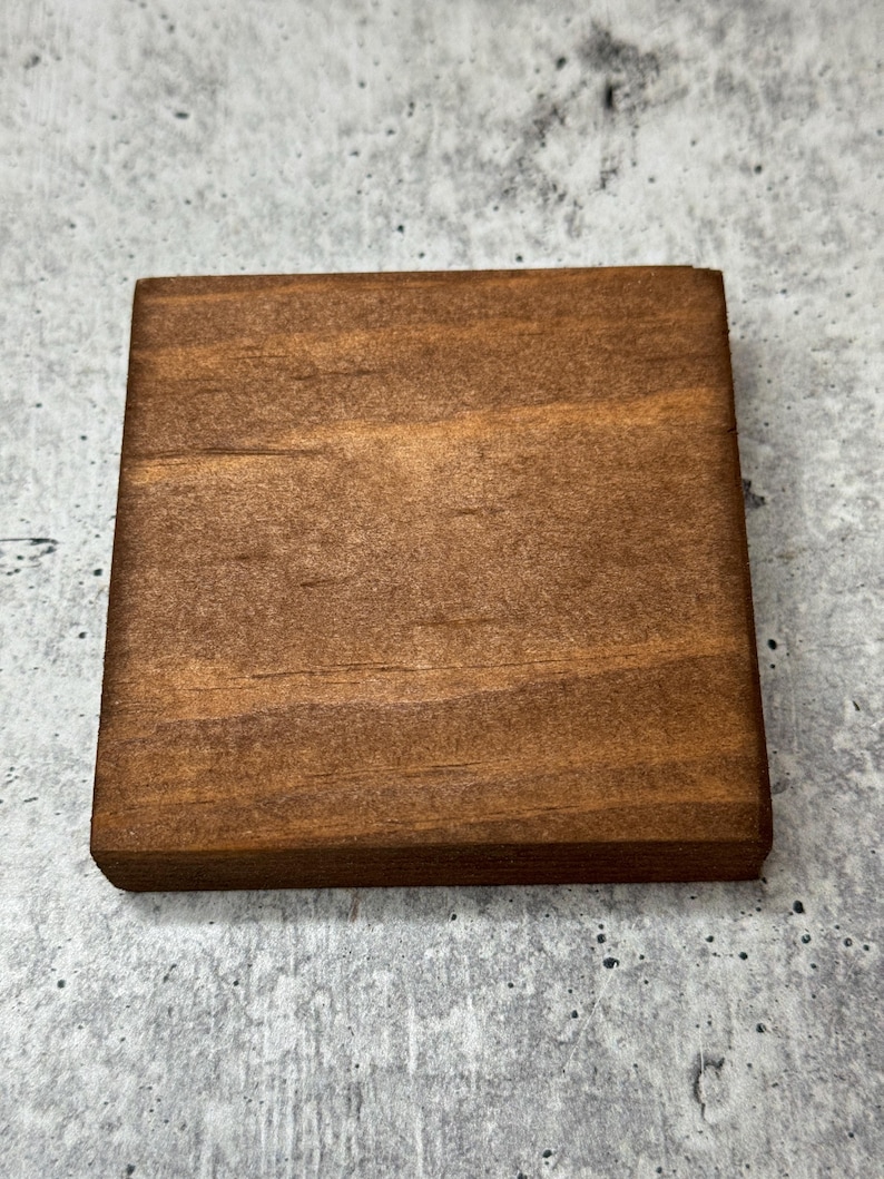 May include: A square wooden coaster with a dark brown stain. The wood grain is visible.