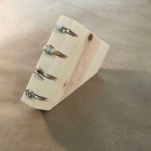 May include: A wooden ring holder with five slots for rings. The holder is made of light wood and has a triangular shape.