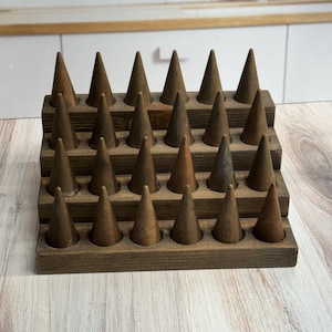 May include: A dark brown wooden ring display stand featuring four tiered rows. Each row contains six upright cone-shaped holders, totaling twenty-four individual wooden cones designed for organizing and showcasing rings in a structured arrangement.