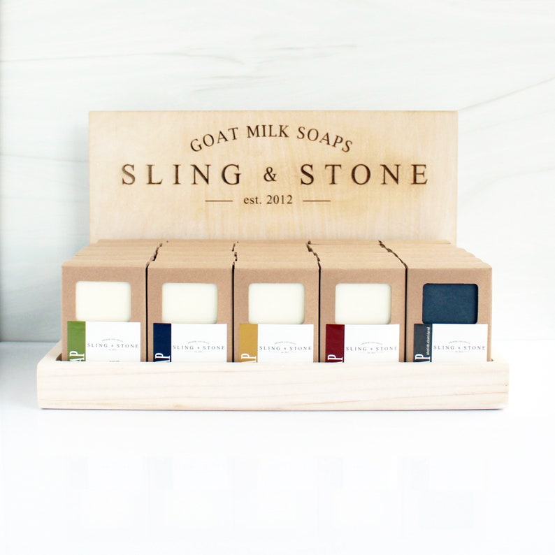 May include: A wooden display stand with a sign that reads 'GOAT MILK SOAPS SLING & STONE est. 2012'. The stand holds multiple boxes of goat milk soap, each with a different color and a label that reads 'SLING & STONE'.