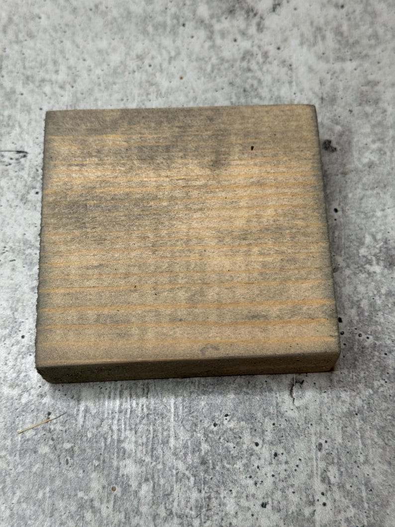 May include: A square piece of wood with a natural wood grain and a light brown color. The wood has a distressed look with some darker areas.