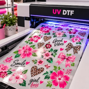 May include: A UV DTF printer outputting a clear film sheet featuring a variety of printed designs including bright pink hibiscus flowers, green leaves, leopard print hearts, pink bows, and the text Good Vibes, Smile, and girl boss in black script.